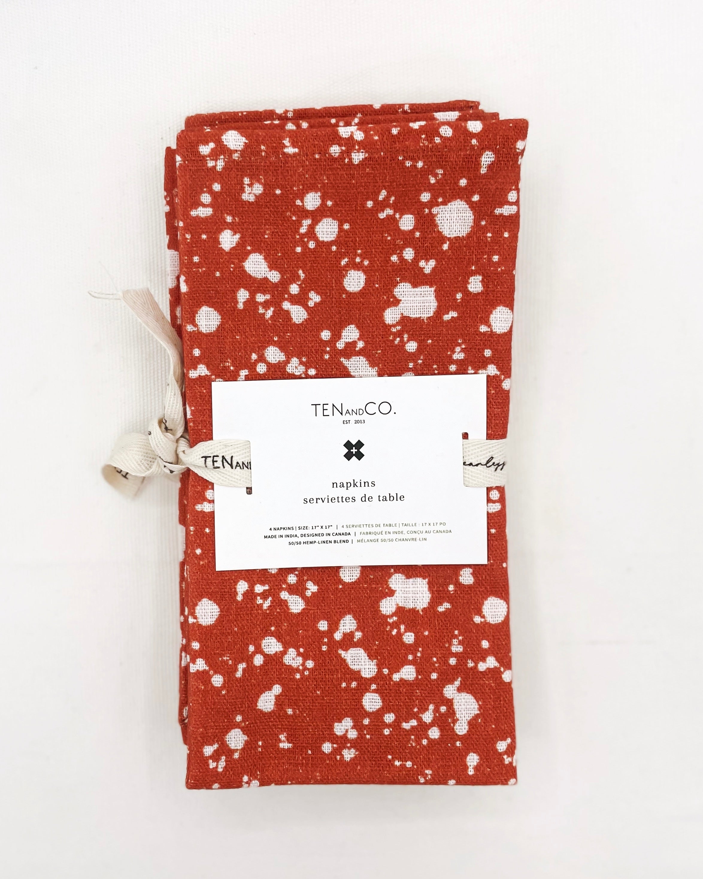 Flat lay image of Everyday Napkins Set of 4, Splatter – Rust on a white background. The napkins have a deep orange base colour with splatters of white covering the entirety of the cloth and are tied together with Ten and Co twill tape and a recyclable paper tag laying on top.