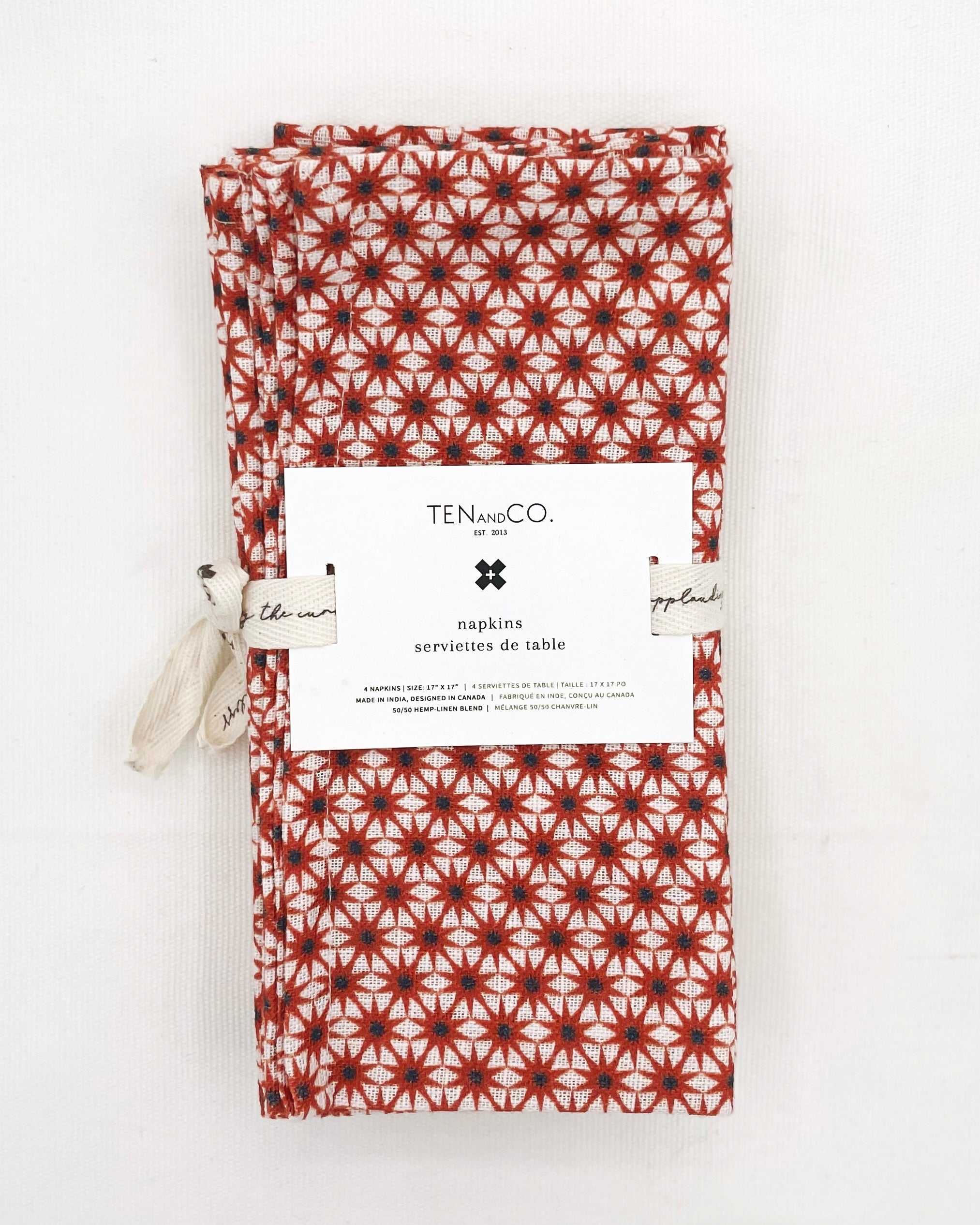 Flat lay image of Starburst Rust, 4 set of Napkins on a white background. The napkins have a white base colour with starbursts of red covering the entirety of the cloth and are tied together with Ten and Co twill tape and a recyclable paper tag laying on top.