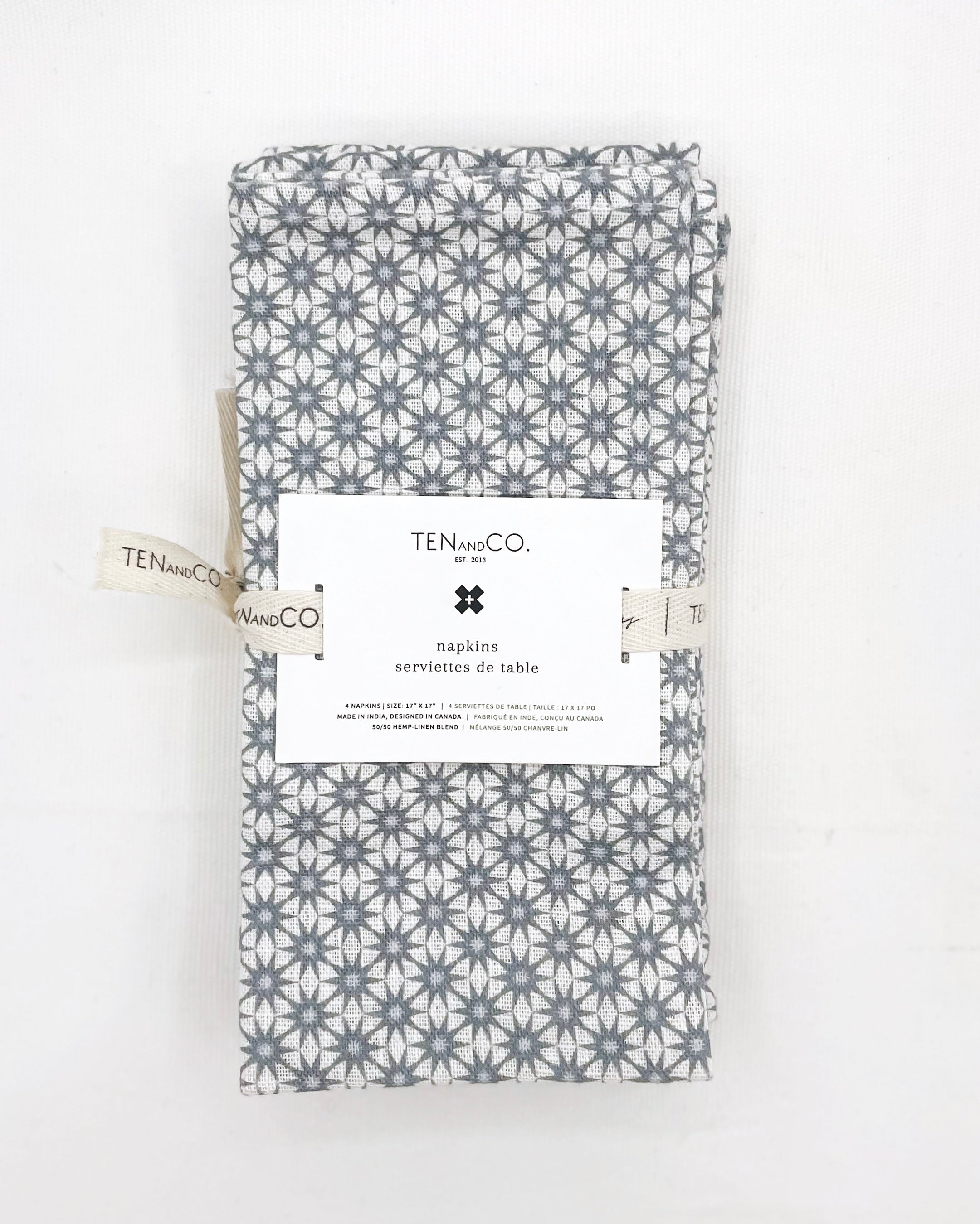 Flat lay image of Starbust Denim, 4 set of Napkins on a white background. The napkins have a white base colour with starbursts of blue covering the entirety of the cloth and are tied together with Ten and Co twill tape and a recyclable paper tag laying on top.
