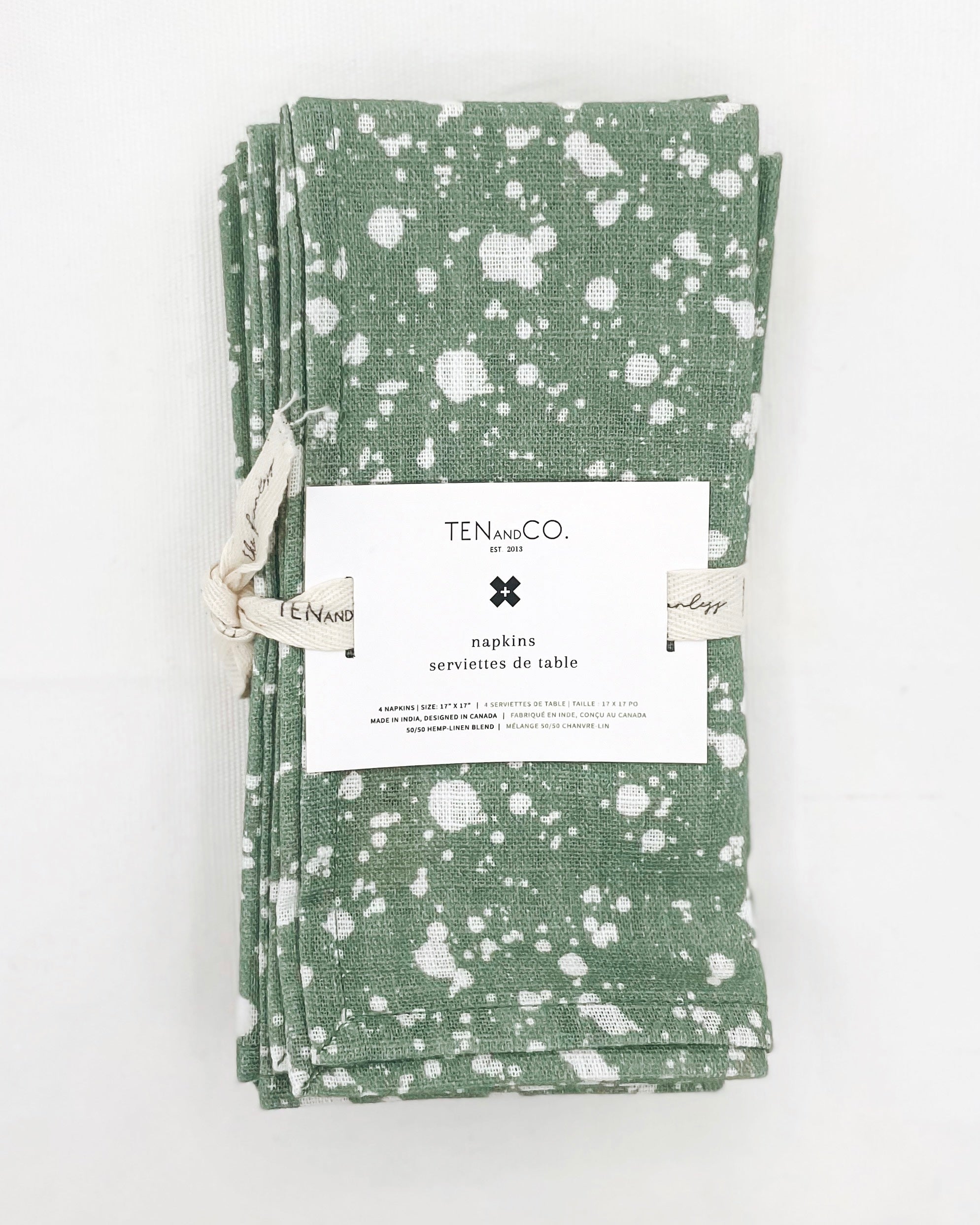 Flat lay image of Everyday Napkins Set of 4, Splatter Sage on a white background. The napkins have a medium green base colour with splatters of white covering the entirety of the cloth and are tied together with Ten and Co twill tape and a recyclable paper tag laying on top.