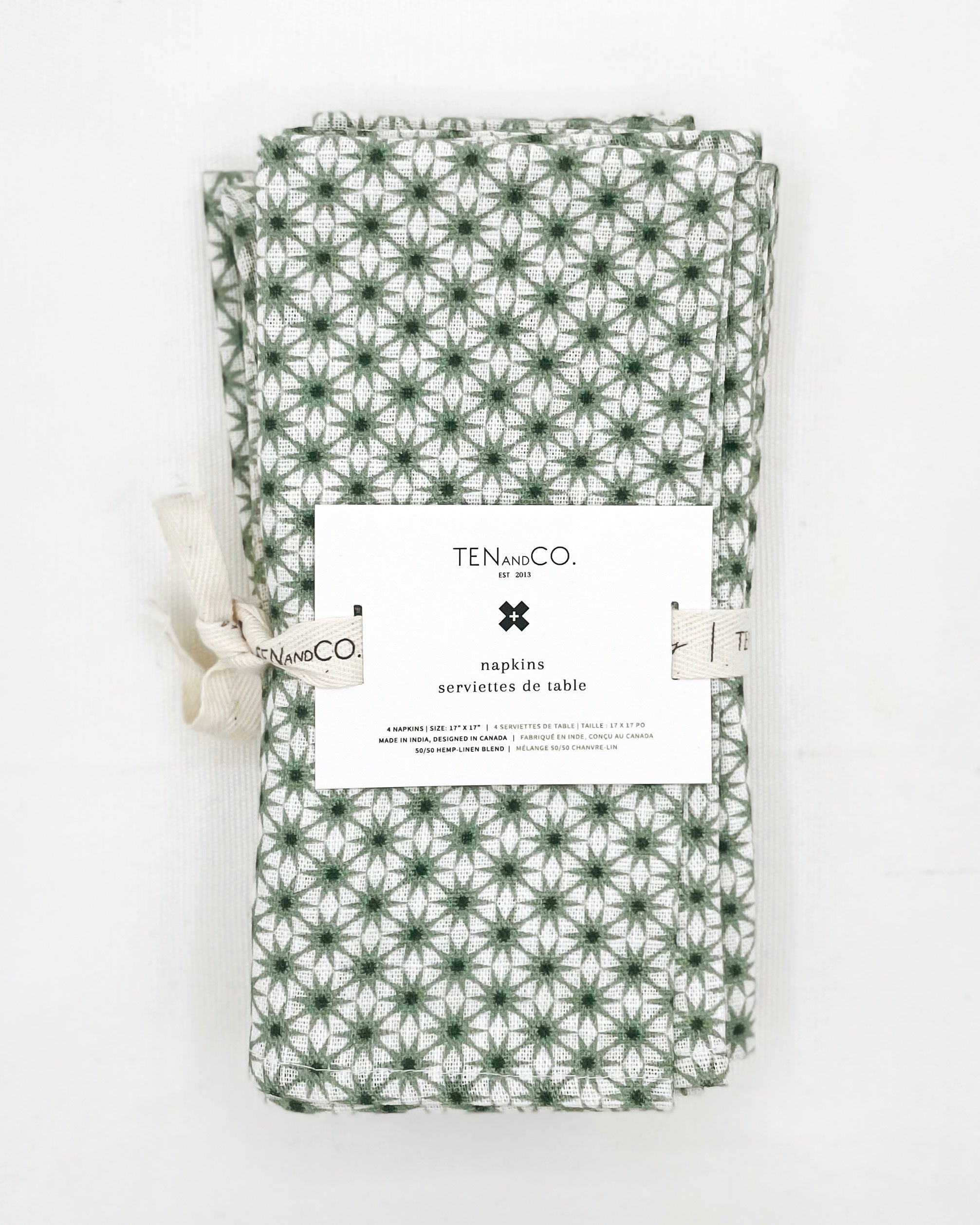 Flat lay image of Starburst Sage, 4 set of Napkins on a white background. The napkins have a white base colour with starbursts of green covering the entirety of the cloth and are tied together with Ten and Co twill tape and a recyclable paper tag laying on top.