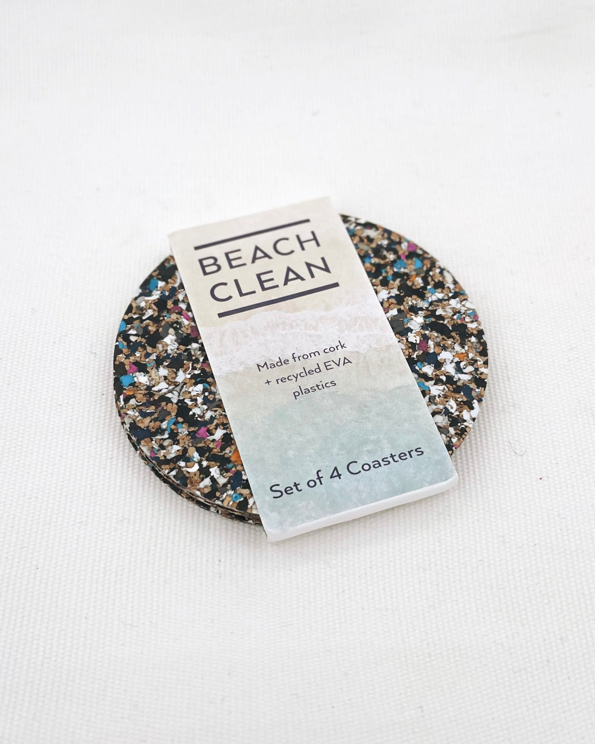 Product image of Beach Clean Coasters on a white background. The product is on an angle. They are round and multi-coloured cork. There is a belly band around the coasters vertically. It is pink and blue with the Beach Clean logo in blue font along with descriptive text.