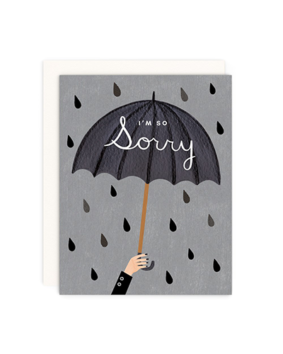 Flat lay image of I'm So Sorry Card on a white background. The card has a grey base with black and grey raindrops throughout. There is a hand coming out of the bottom of the card holding an open umbrella. The umbrella is dark grey with “I’m So Sorry” written in white font. There is a white envelope behind the card.