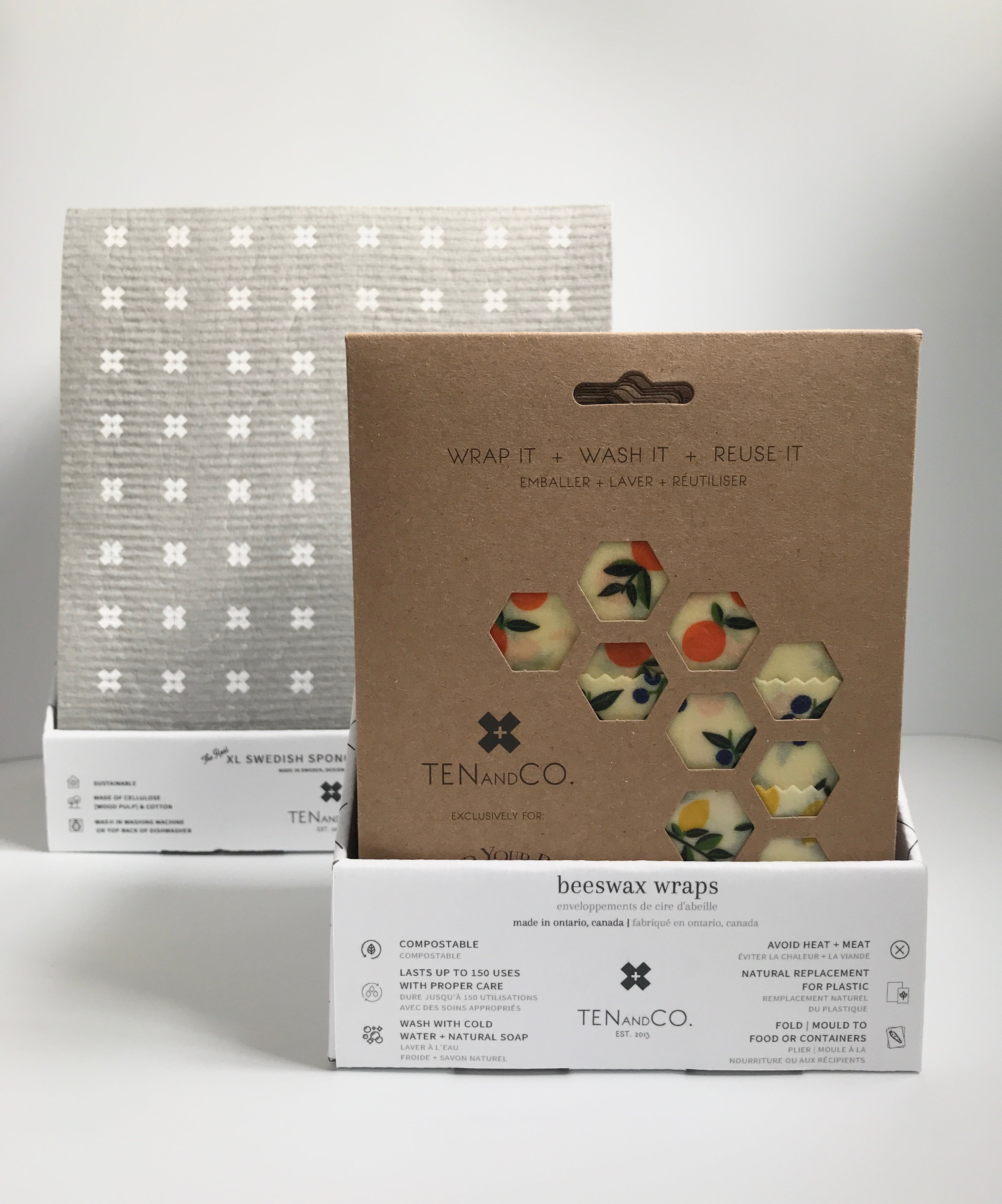  Straight on image of the Cardboard Display Box for Beeswax Wraps and Cardboard Display Box for Sponge Cloths. Both boxes are on a white surface with white background. The Beeswax Wraps display is on the right side and a little bit closer, whereas the Sponge Cloth display is on the left side and a little bit farther back. The Beeswax Wraps package at the front of the display box is Vintage Fruits and the Sponge Cloth at the front of the display box is Tiny X White on Grey. 