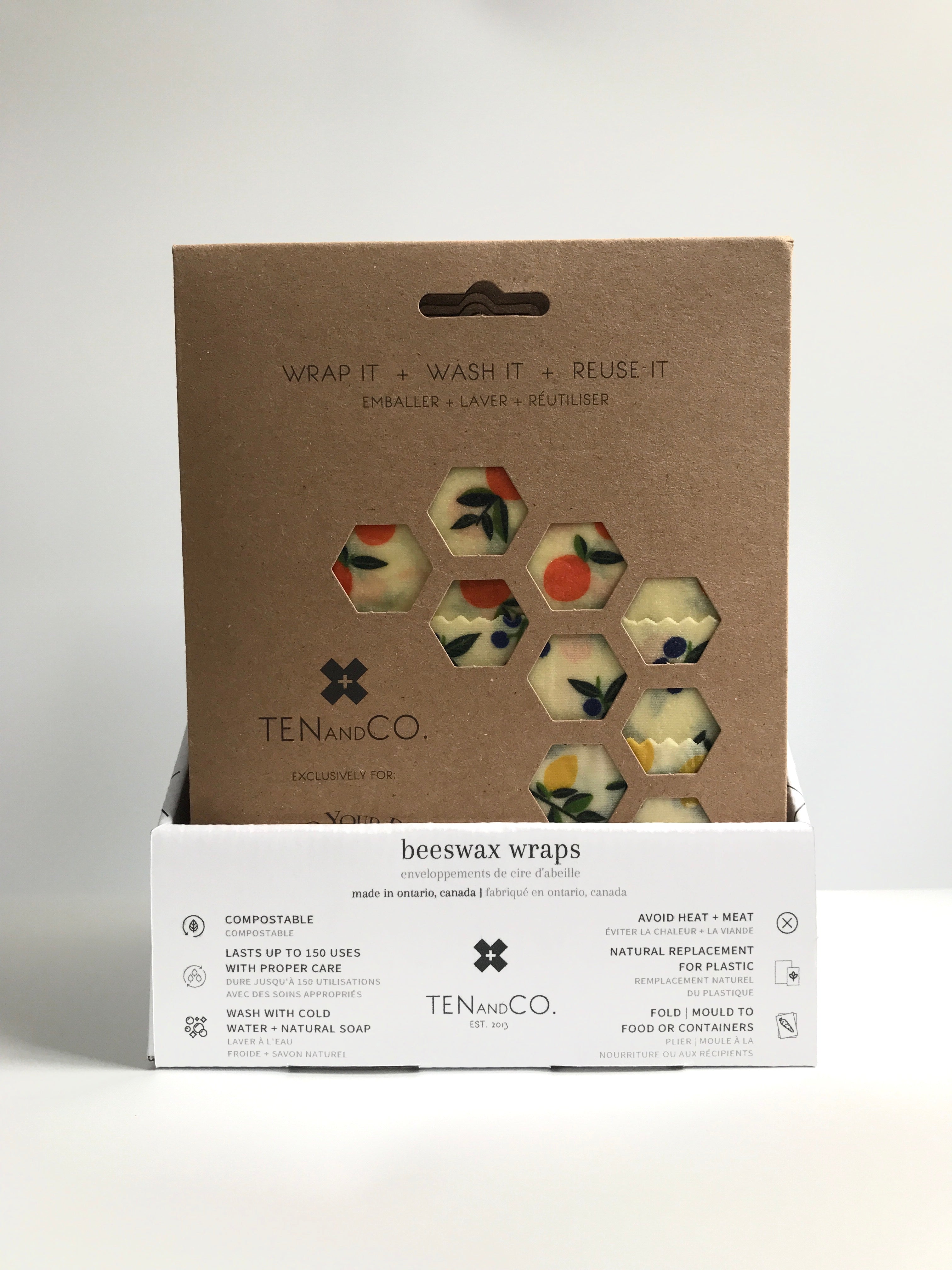 Straight on image of the Cardboard Display Box for Beeswax Wraps. Box is on a white surface and white background. The box is white with black detailing. The front of the box has the Ten and Co logo in the middle and descriptions of Beeswax wraps in black. The box is filled with Beeswax wraps in brown packaging. The front package is Vintage Fruits, which can be seen through a few hexagonal holes in the packaging. 