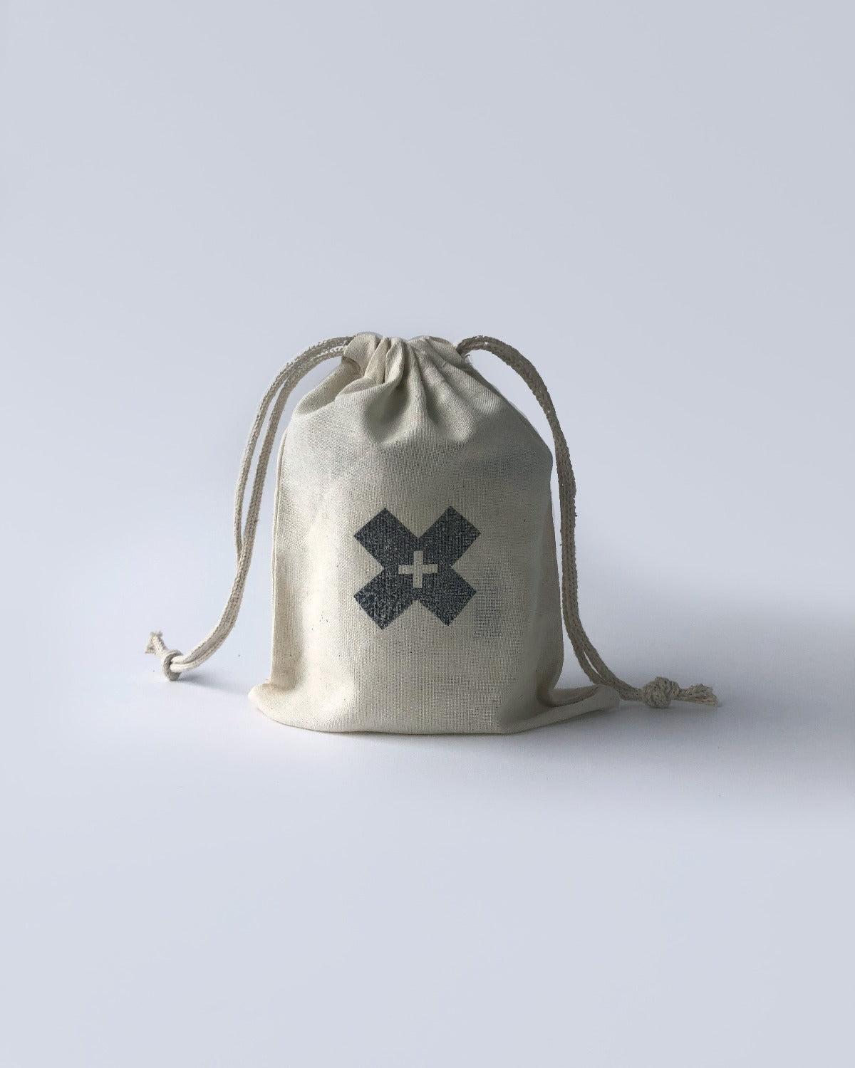 Product image of Reusable Cotton Drawstring Bag. The bag is a beige cotton with a large black X in the middle with a tiny cross in the X. There are strings coming out of either side of the top and the bag is closed tight. 