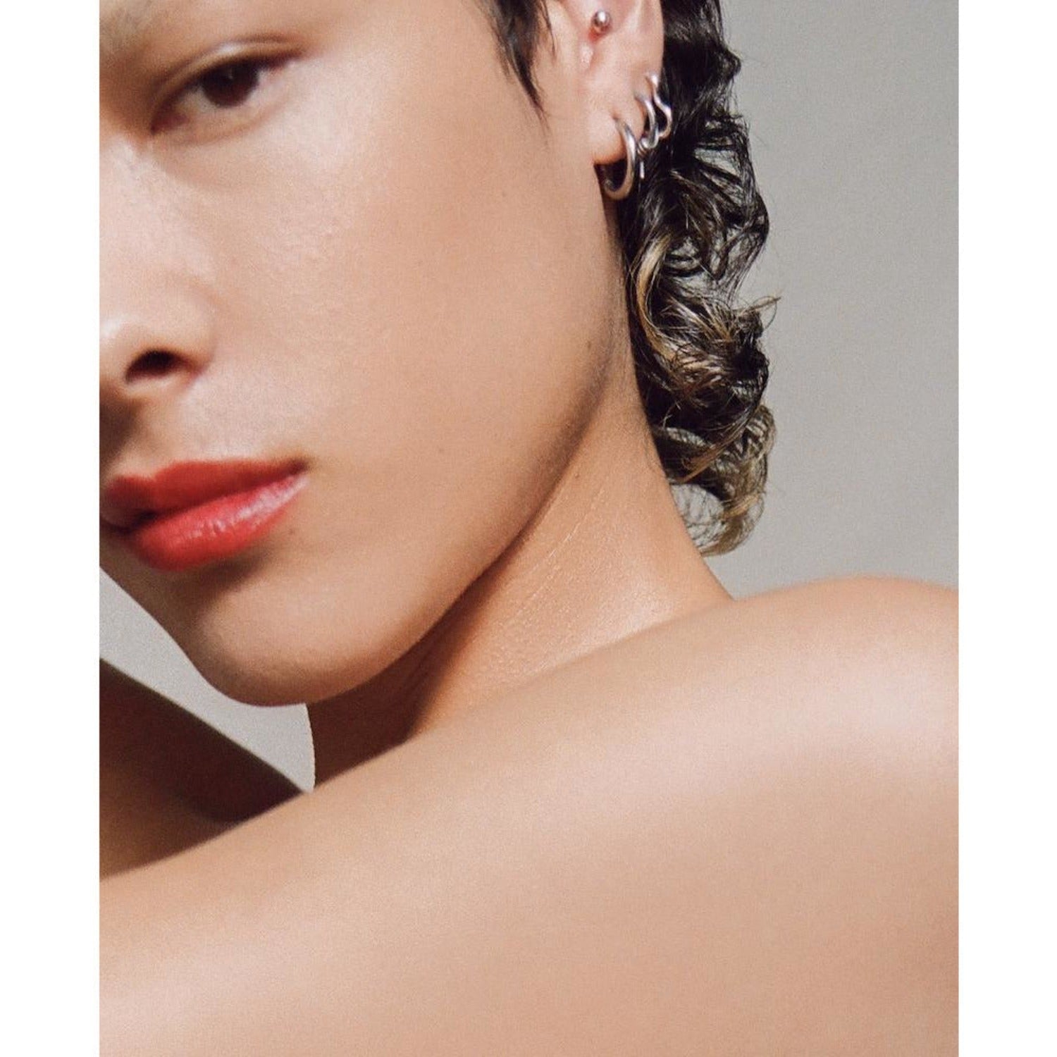Product image of Multi Bene Stain Stick Oscillate on a white background. There is a side profile of a person with short curly hair. They have many earrings on. There arm and shoulder is taking up the lower half of the image. They have pink lips. 
