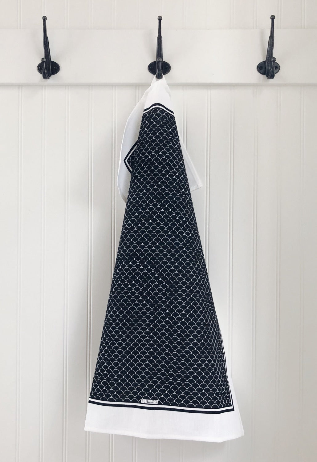 Product image of Scallop Black gift set. In this image there are 3 black hooks on a white wood paneled wall. On the center hook is a full view of the Scallop Black tea towel. It has a white base with a black background and white scallops throughout. 