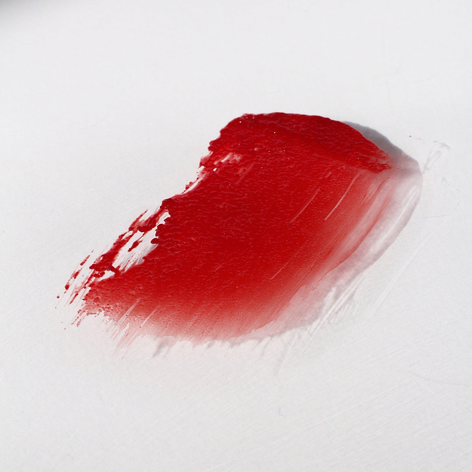 Flat lay image of Multi Bene Stain Stick Oscillate on a white background. The product has been smeared on a white surface so you can see the colour. It is a pinky red shade and is shiny. 