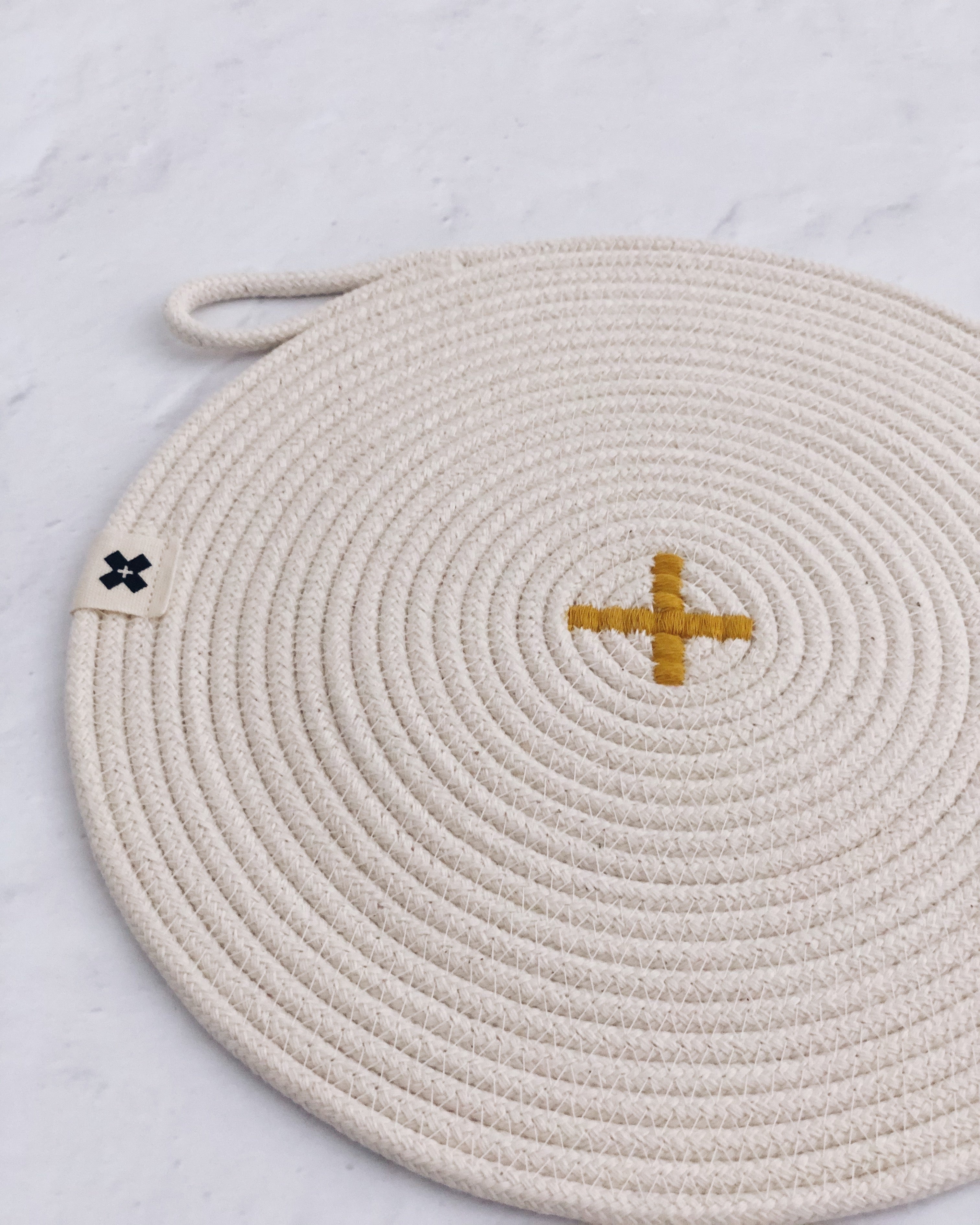 Close up image of Rope Trivet Ochre. The white rope trivet is laying on a white surface. The trivet is a white cotton rope spiral with a yellow X in the middle. There is a tiny Ten and Co. Logo attached to one of the sides. There is a loop at the end of the rope to use for hanging.
