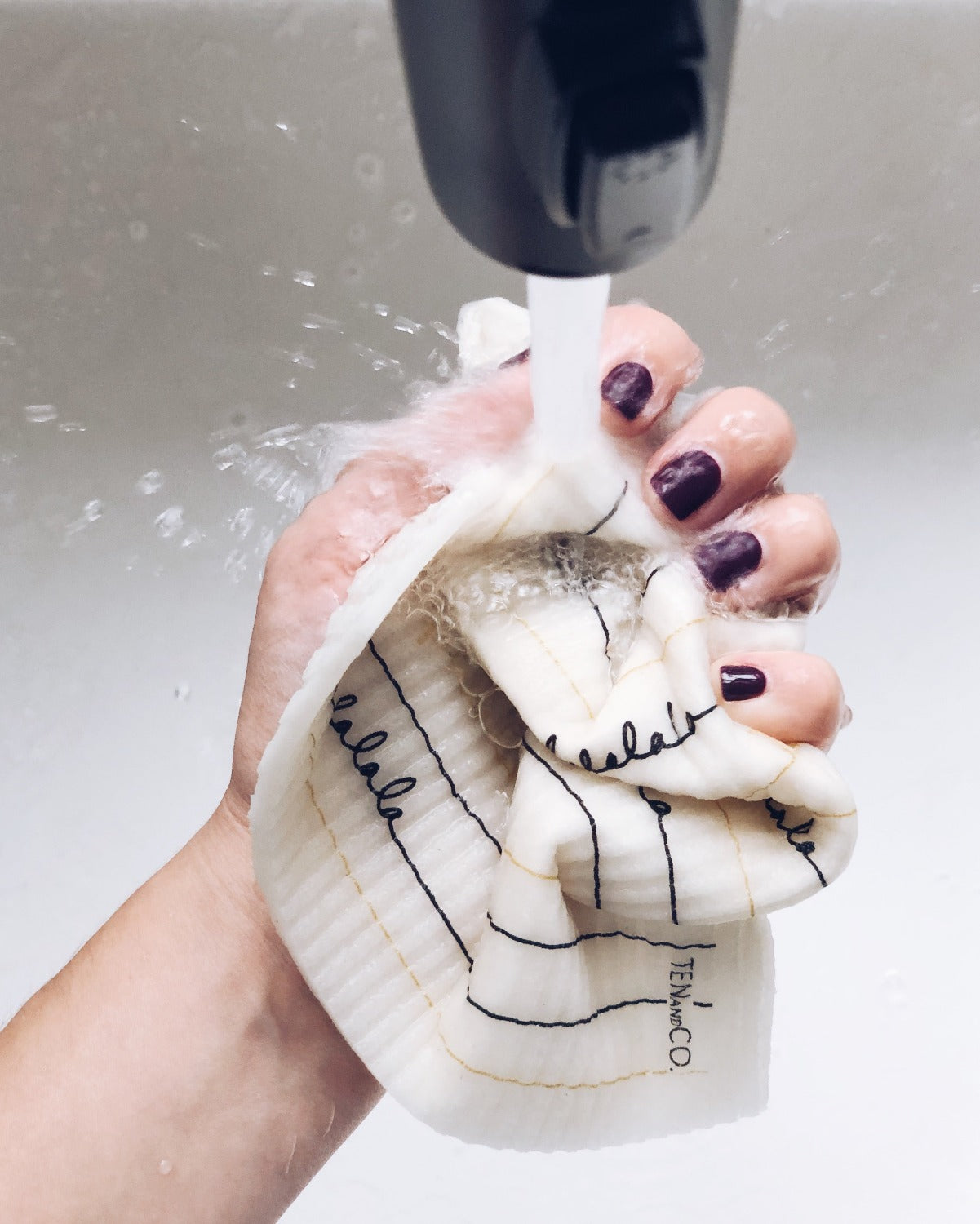 Product image of Falala in Black and Gold in a white sink. There is a black faucet coming out of the top of the image. There is water coming out of the faucet onto a hand holding the Falala Black and Gold sponge cloth. The hand is squeezing a cloth into a ball. The hand has purple nail polish on the nails. 